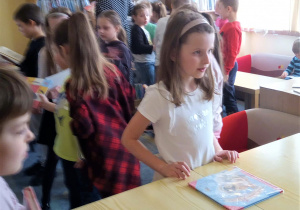 Dwie uczennice stoją przy stole i wypożyczają książki. W tle grupa dzieci stojąca przy regale z książkami.